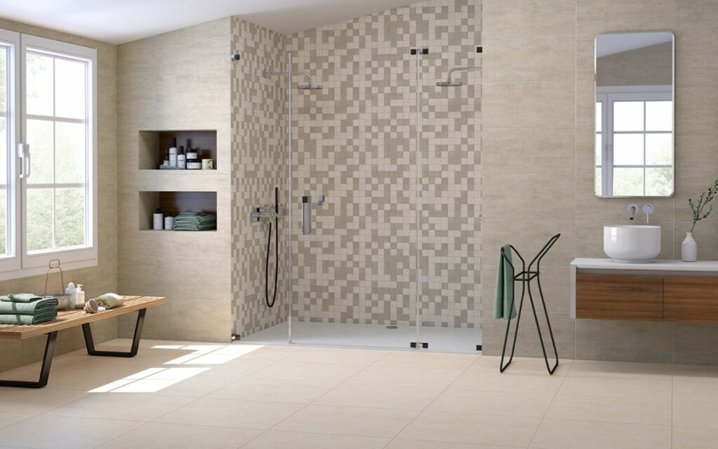 Amplia sala de baño con pavimento y revestimiento efecto piedra en tono beige. Zona de ducha alicatada con cerámica tipo mosaico. Plato de ducha blanco rectangular. Ducha con dos rociadores y grifería acabado cromo. Mampara de puerta con bisagra. Lavabo sobre encimera de fondo profundo y grifería mural monomando con caño y mando separados en acabado cromo. Mueble de baño suspendido con puertas en acabado madera y superficie blanca. Espejo de baño rectangular.