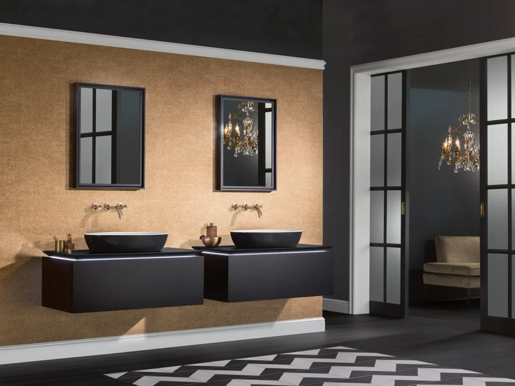 Sala de baño en tonos marrones con dos muebles suspendidos, independientes con lavabo negro sobre encimera cada uno. En la pared, revestida con cerámica efecto metal en tono dorado, grifería empotrada en color oro y acabado cepillado y dos espejos rectangulares con marco en negro. El suelo en cerámica oscura forma en el centro una bonita alfombra porcelánica con piezas blancas y negras colocadas en espiga