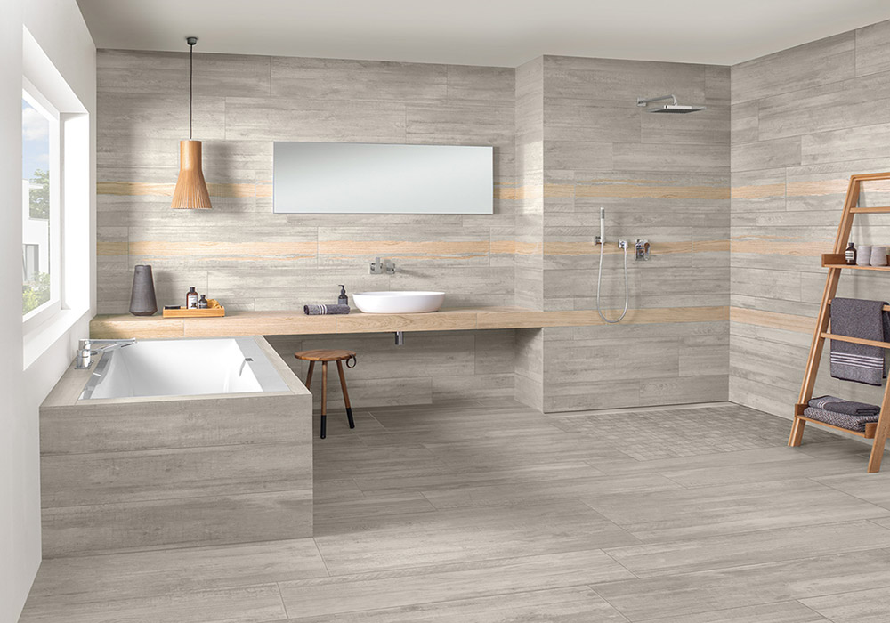 Fantástico cuarto de baño revestido y pavimentado con cerámica efecto madera en tonos grises y detalle decorativo en marrón en la zona de la encimera sobre la que se coloca el lavabo de forma oval y color blanco. La bañera también está revestida con la misma cerámica en tono gris